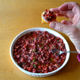 A hand holding a cracker with cranberry cream cheese dip on it with a casserole dish of dip below it. 