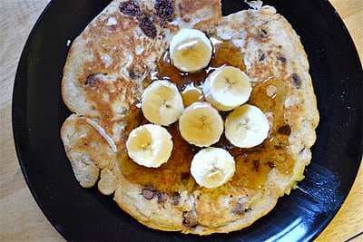 A chocolate chip pancake topped with bananas and maple syrup on a black plate. 