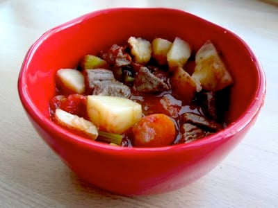 A bowl of beef stew with potatoes, carrots and celery in a red bowl. 