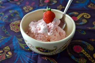 A bowl of strawberry ice cream with a spoon and fresh strawberry on top on a floral table cloth.